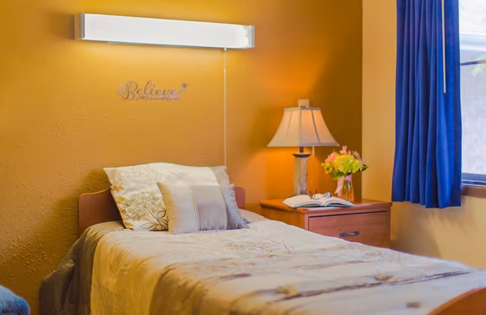 A cozy bedroom with a single bed made up with beige and white bedding and pillows. A wooden nightstand beside the bed holds a table lamp, an open book, and a vase with flowers. A window with blue curtains is on the right side, and a wall light is mounted above the bed. The wall behind the bed is painted a warm yellow color with the word 'Believe' displayed on it.