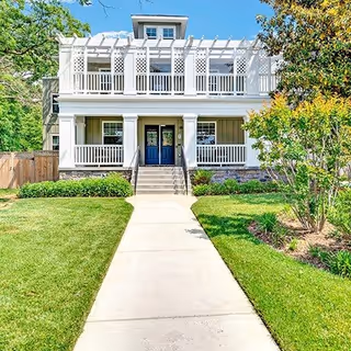 Front exterior view of a two-story white building with a porch and balcony, surrounded by green grass, trees, and shrubs under a clear blue sky.
