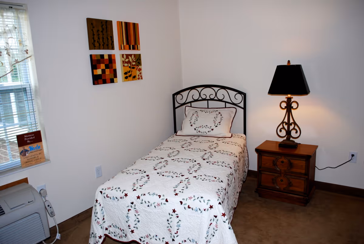 A small bedroom with a single bed covered with a white quilt featuring a floral wreath pattern. Next to the bed is a wooden nightstand with two drawers and an ornate lamp with a black lampshade. On the wall above the bed are four square decorative art pieces with abstract and floral designs. A window with blinds is partially visible on the left side, and an air conditioning unit is below the window.