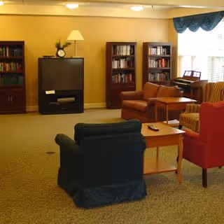 Cozy common room with armchairs, side tables, bookshelves, a TV and a piano near a window.