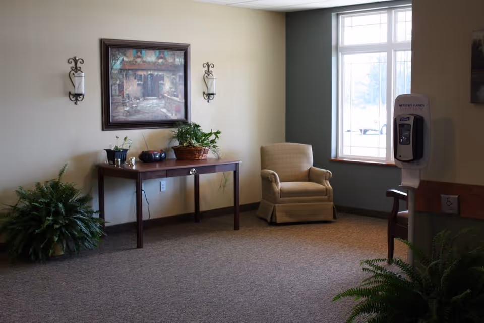 A quiet sitting area in a memory care facility featuring a beige armchair near a large window, a wooden table with potted plants and a small radio, wall-mounted candle holders, framed artwork above the table, and a hand sanitizer dispenser mounted on a stand near the window. The room has carpeted flooring and neutral-colored walls.