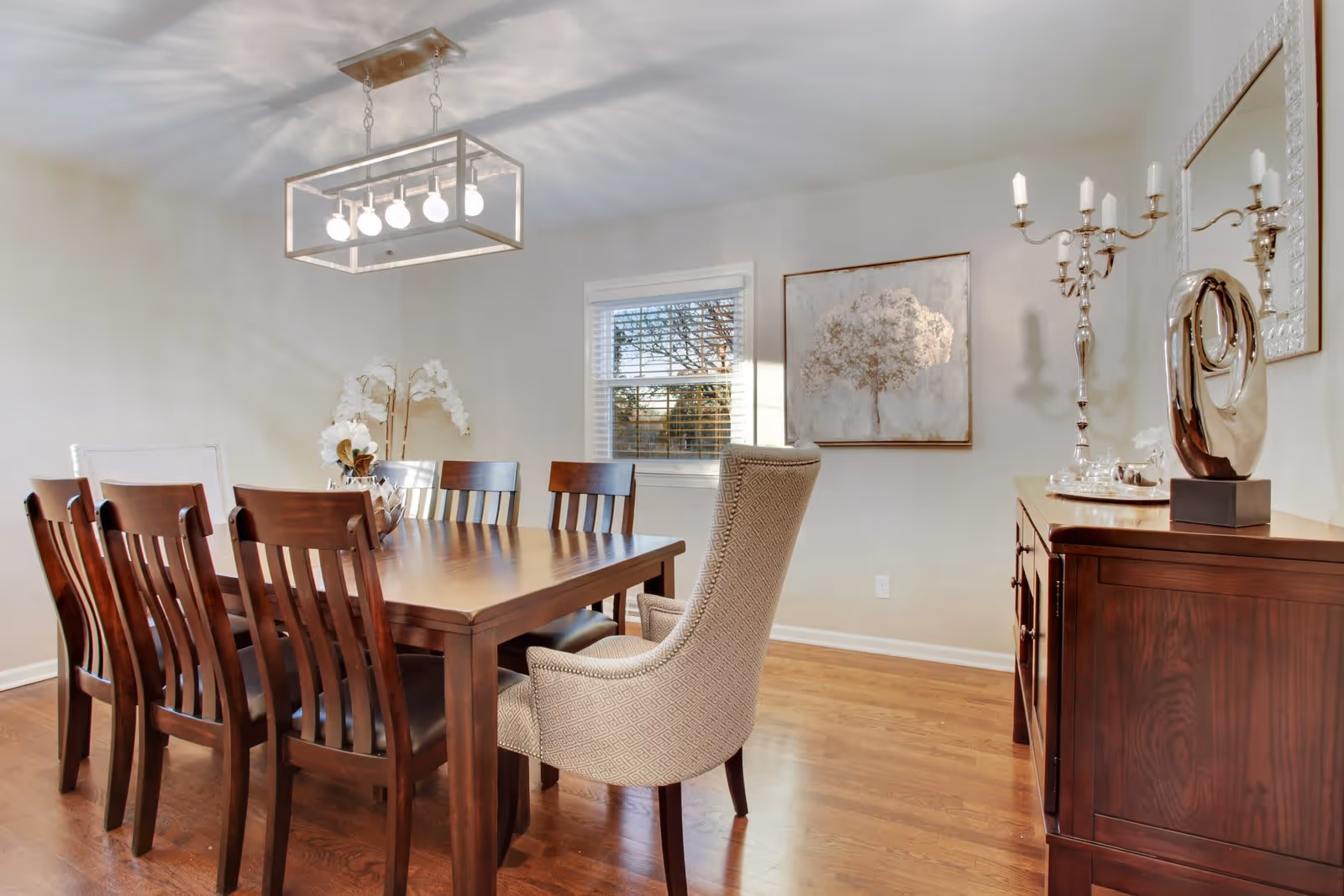 A well-lit dining room with a wooden dining table surrounded by eight chairs, including a cushioned armchair at the head of the table. The room features hardwood flooring, a modern rectangular chandelier with exposed bulbs hanging above the table, a window with white blinds, a painting of a tree on the wall, and a wooden sideboard with decorative items including a silver candelabra and a modern sculpture.
