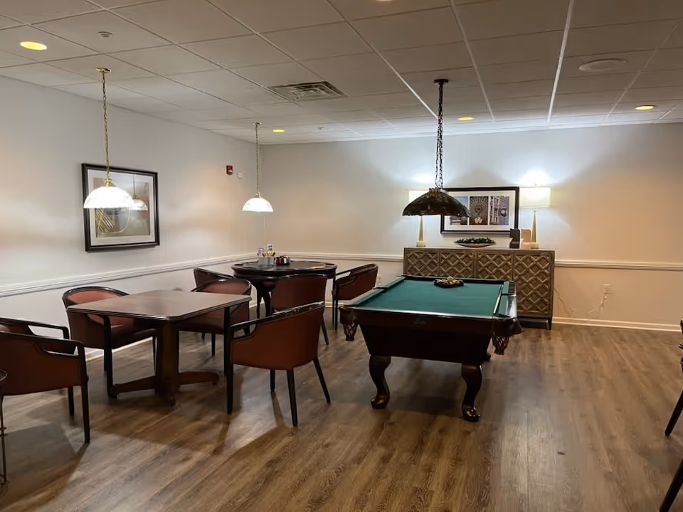 A common area with a pool table in the center and two dining tables with chairs around them. The room has wood flooring, white walls with a chair rail, and ceiling lights. A sideboard with two lamps and framed artwork is visible against the far wall.