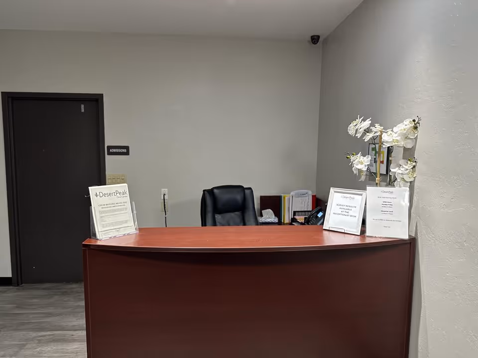 Reception desk area at Desert Peak Care Center with a dark wooden counter, a black office chair behind it, and informational signs on the desk. There is a door labeled 'Admissions' to the left and a vase with white flowers on the right side of the desk.