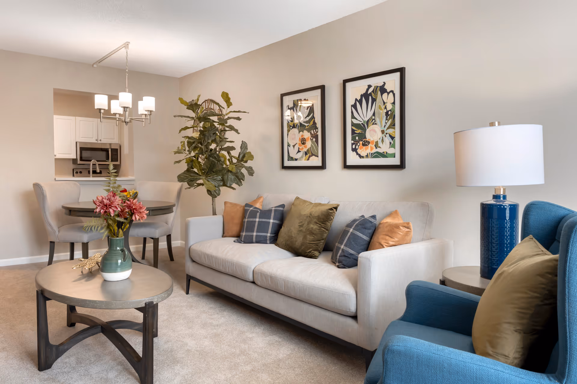 Well-lit living room with a sofa, blue armchair, coffee table, dining nook and kitchen pass-through, decorated with framed artwork and a potted plant.
