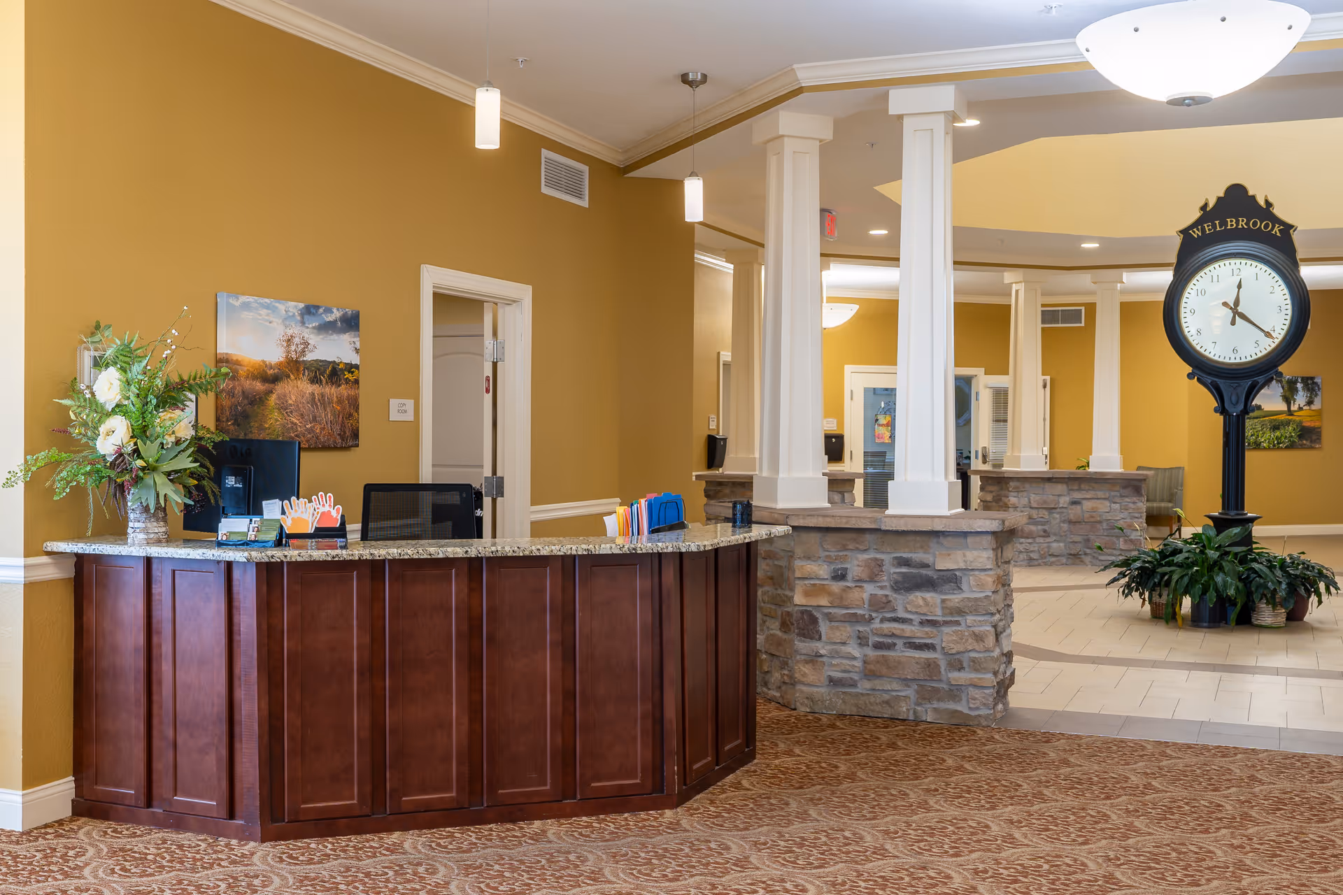 Reception area with a wooden front desk, a computer, and a flower arrangement on the left. Behind the desk is a wall with a landscape painting. The area has beige walls, carpeted floor, and stone pillars. In the background, there is a large black clock with the word 'WELBROOK' on it, surrounded by plants.