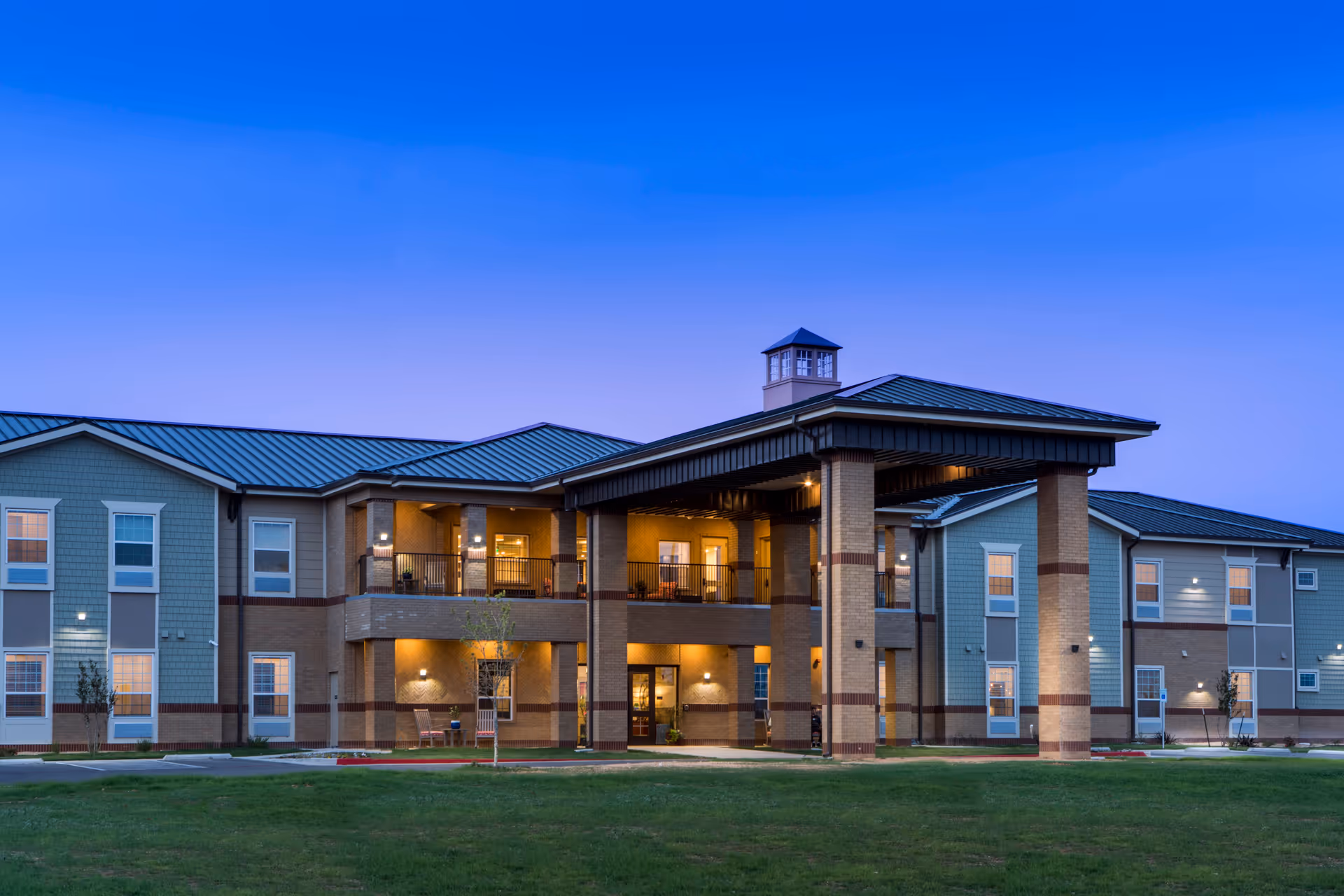 Exterior view of a two-story senior living facility building at dusk with lights on inside. The building has a covered entrance supported by large pillars, multiple windows, and a green lawn in front under a clear blue sky.