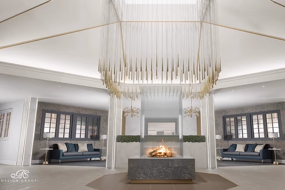 A modern, elegant living room area featuring a central glass-enclosed fireplace with a fire burning inside. Above the fireplace hangs a large, artistic chandelier with numerous thin, vertical gold rods. On either side of the fireplace are two seating areas with dark blue sofas, side tables, and lamps. The walls have large windows with dark frames, and the overall color scheme is neutral with gold accents.