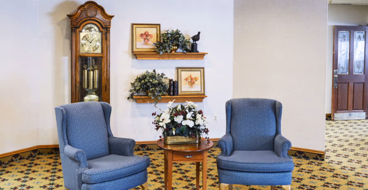 Two blue upholstered armchairs flank a small wooden table with a floral arrangement in a decorated lounge area featuring a grandfather clock and wall shelves.