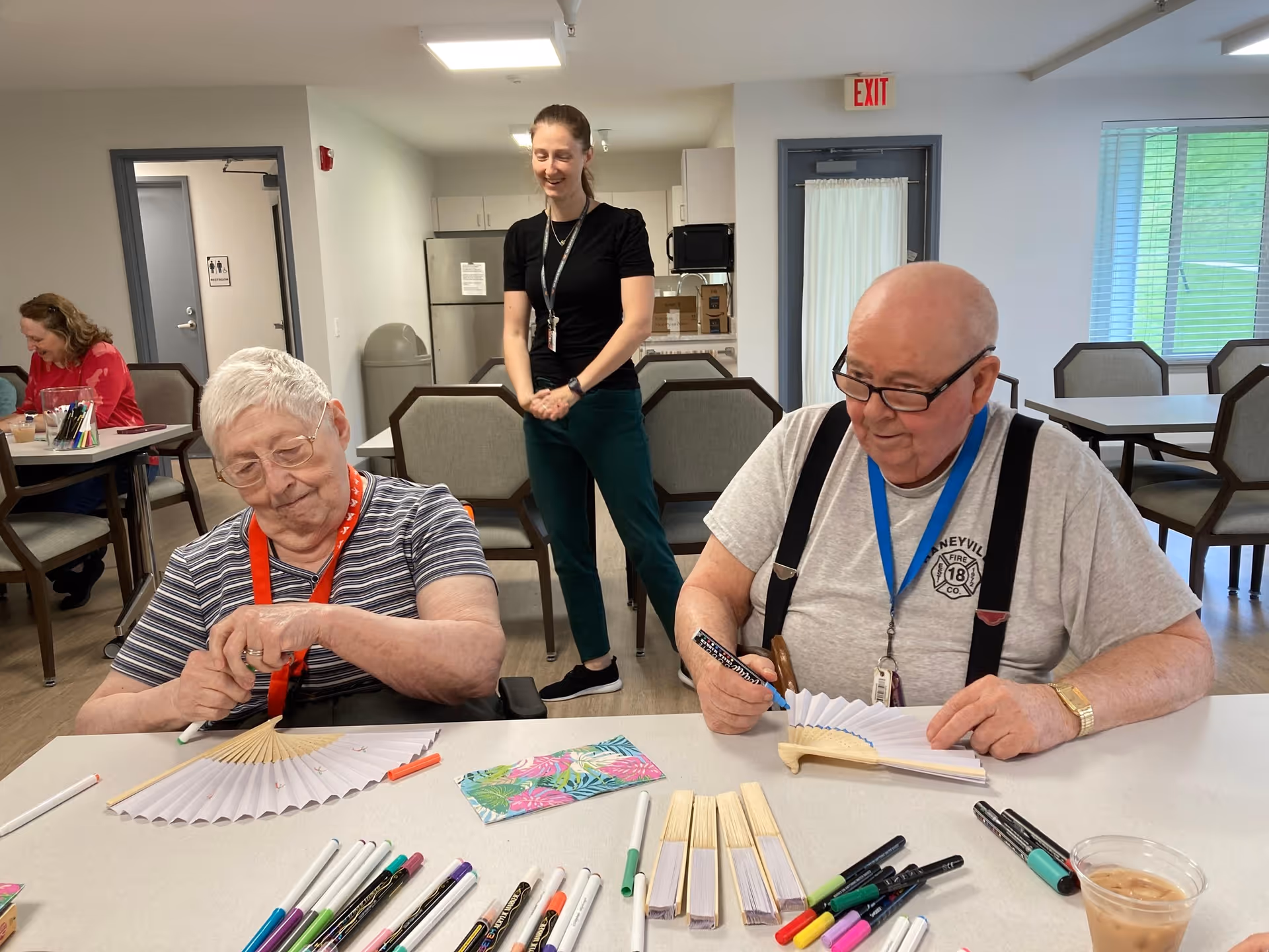 Two seniors seated at a table decorating paper hand fans while a staff member stands behind them in a communal activity room.
