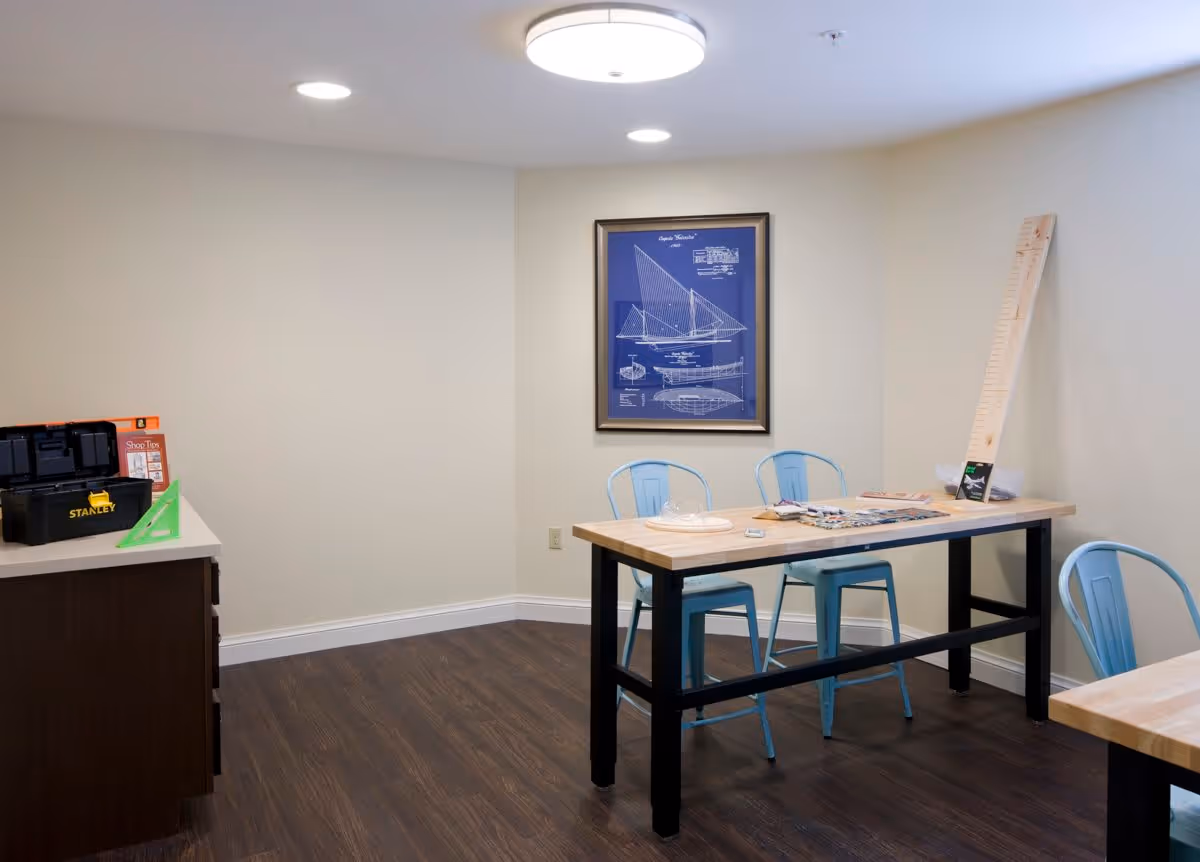 A room with wooden flooring and cream-colored walls featuring a wooden table with black legs and three light blue metal chairs. On the table are various items including a wooden ruler, papers, and a small box. A framed blueprint of a sailboat hangs on the wall. To the left, there is a countertop with a Stanley toolbox and other items.