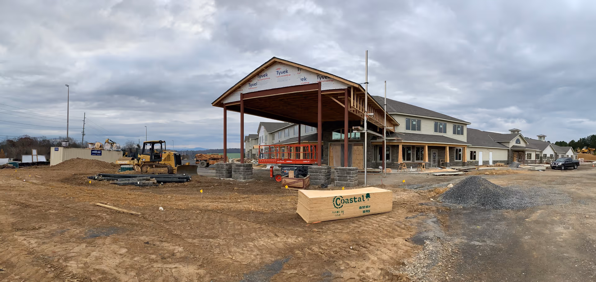 Construction site of a large building with exposed framing and construction materials scattered around. The building appears to be a senior living facility under construction with a covered entrance area and multiple windows. The sky is cloudy and the ground is mostly dirt and gravel.
