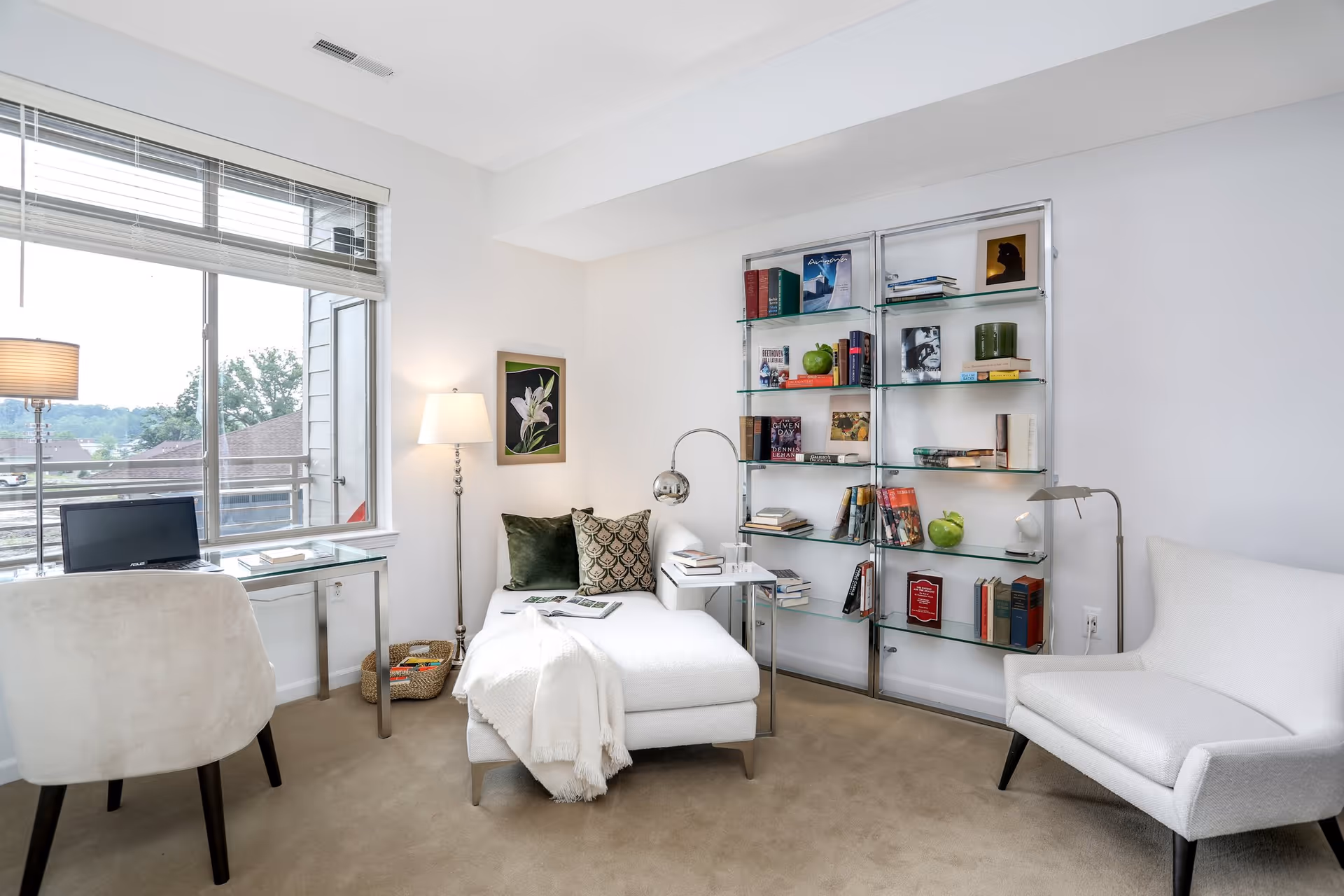Bright modern living area with a chaise lounge, glass bookshelves, a desk by a large window, and two white chairs.