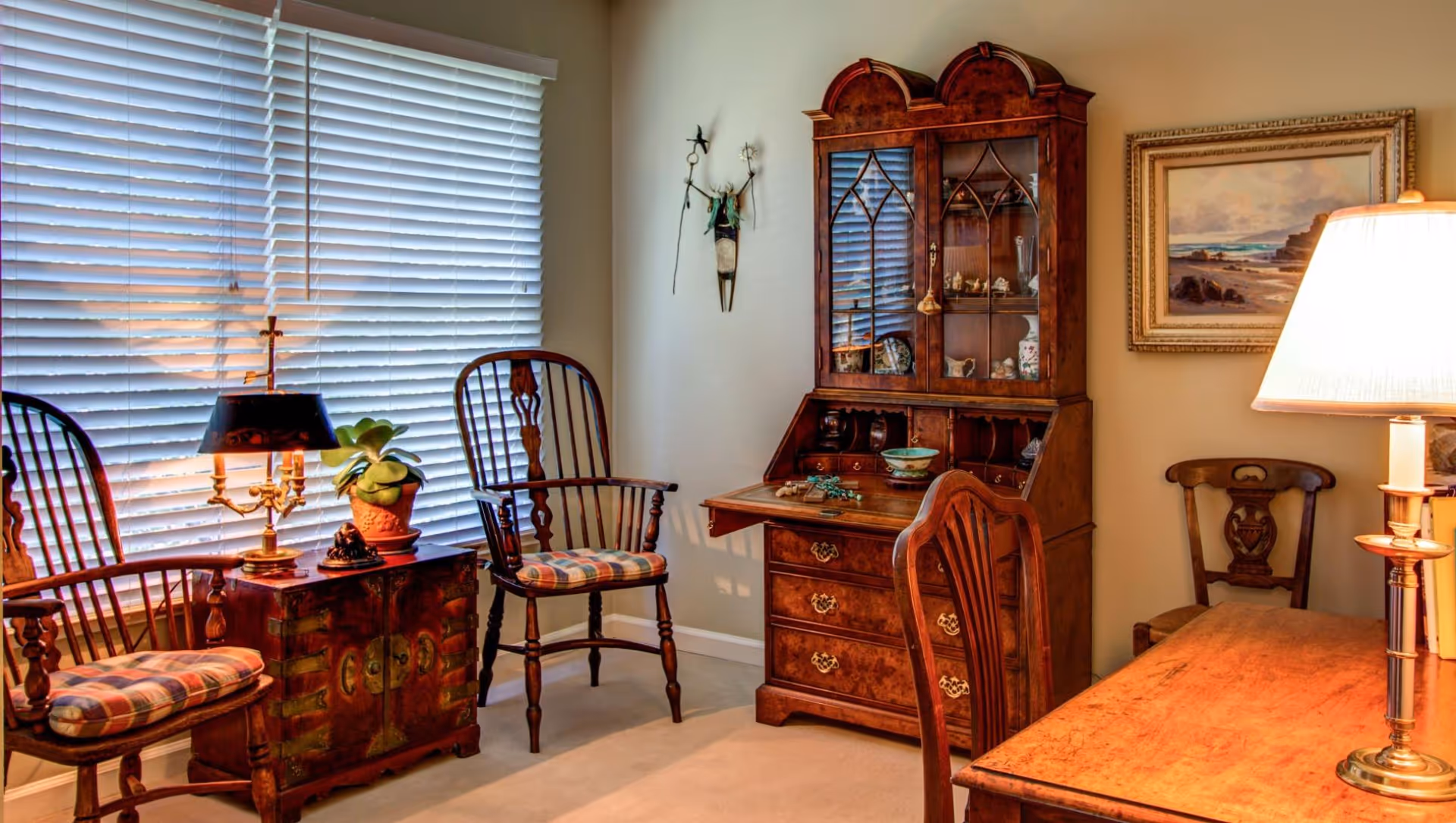 Antique-furnished sitting room with wooden chairs, a writing desk and glass-front hutch beside a large window with blinds.