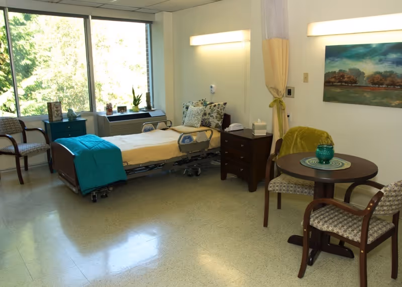 Well-lit senior resident bedroom with a hospital-style bed, bedside table, small dining table and chairs, and a large window overlooking trees.