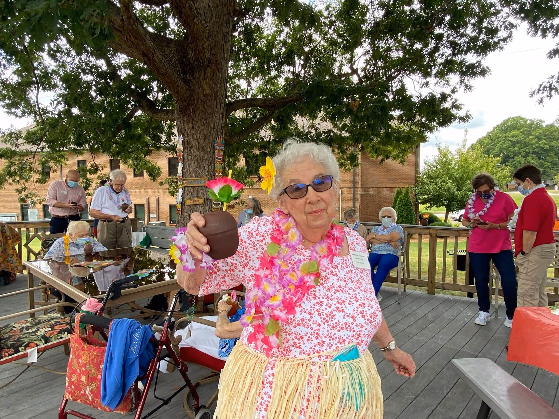 An elderly woman wearing a pink floral shirt, sunglasses, a pink and green lei, and a grass skirt is holding a tropical drink with a flower decoration while standing on a wooden deck. Several other elderly people, some wearing masks, are seated or standing around the deck under a large tree, with a brick building and greenery in the background.