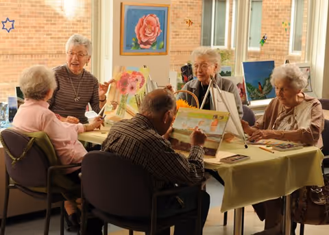 A group of elderly people sitting around a table in a well-lit room, engaged in painting on canvases placed on easels. The room has large windows with a view of a brick wall outside, and colorful artwork is displayed on the walls. The atmosphere appears calm and social.
