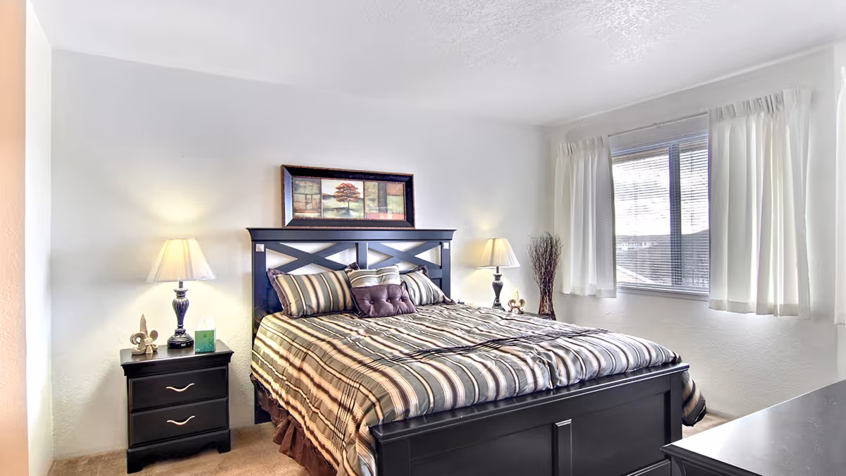 Bright bedroom with a striped bed, dark wooden headboard and matching nightstands with lamps beside a window with white curtains.