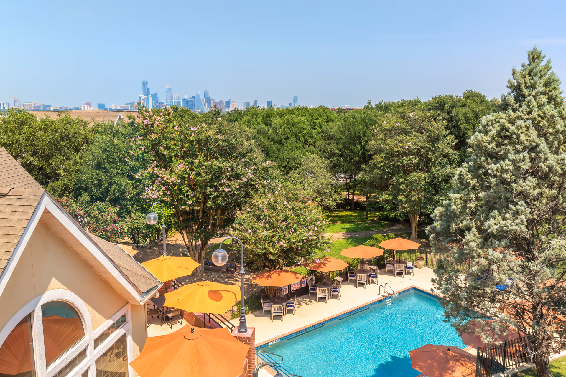 Outdoor swimming pool area with several orange umbrellas and tables surrounded by trees and greenery. A building with a peaked roof and arched windows is visible on the left. In the background, a city skyline is visible under a clear blue sky.