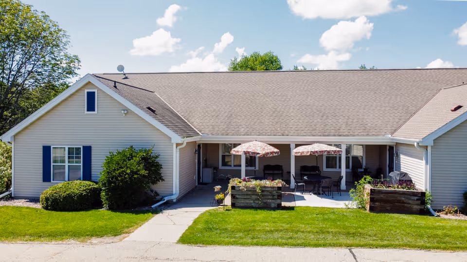 Front exterior of a single-story senior living building with a covered patio, two umbrellas, outdoor seating and a lawn.