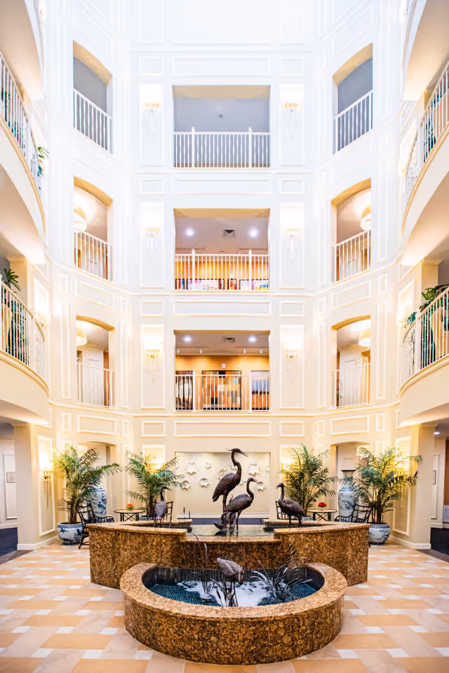 Bright and spacious multi-story atrium with cream-colored walls and railings on each floor. The center features a decorative water fountain with bronze bird sculptures and surrounding plants in large pots. Seating areas with chairs and tables are visible near the fountain.