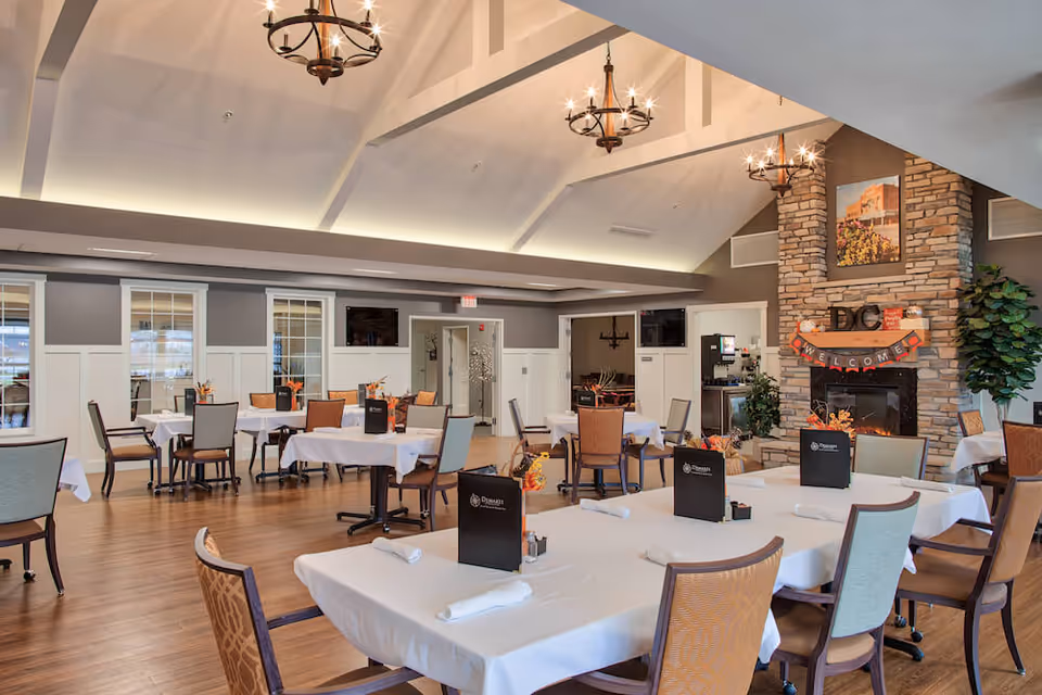 Spacious dining room with multiple tables set with white tablecloths, chandeliers, and a stone fireplace decorated with a welcome banner.
