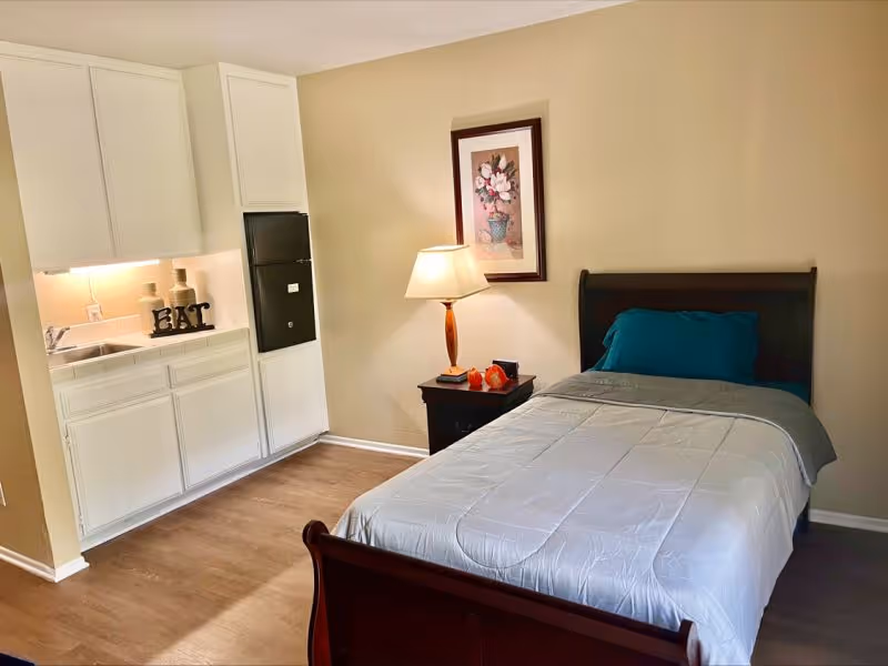 A small bedroom with a single bed made with white and gray bedding and a teal pillow. Next to the bed is a dark wooden nightstand with a lamp, a book, and decorative items. Above the nightstand is a framed floral painting. To the left is a kitchenette area with white cabinets, a sink, and decorative items on the counter, including letters spelling 'EAT'. The floor is wood, and the walls are beige.