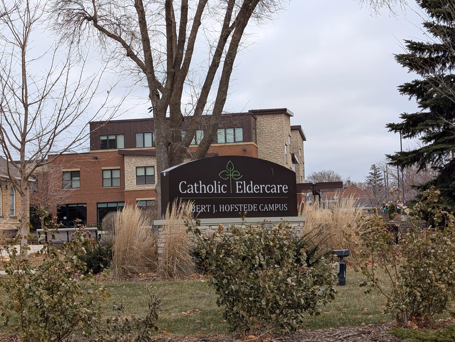 Sign reading "Catholic Eldercare" in front of a multi-story brick and stone eldercare building with shrubs and trees on a landscaped campus.