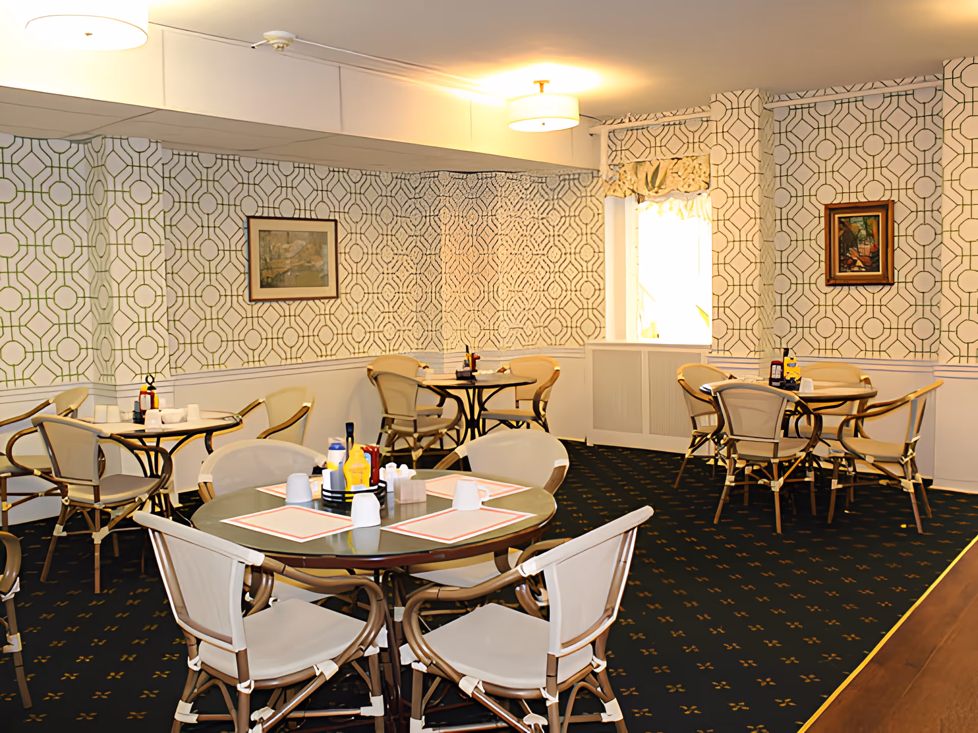 A dining room with several round tables and chairs arranged neatly. Each table has placemats, condiments, and napkins. The walls are decorated with patterned wallpaper and framed artwork. The room is well-lit with ceiling lights and has a window with a valance.