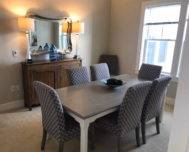 Light-filled dining room with a rectangular wooden table surrounded by six patterned upholstered chairs, a sideboard with a mirror and lamps, and a window.