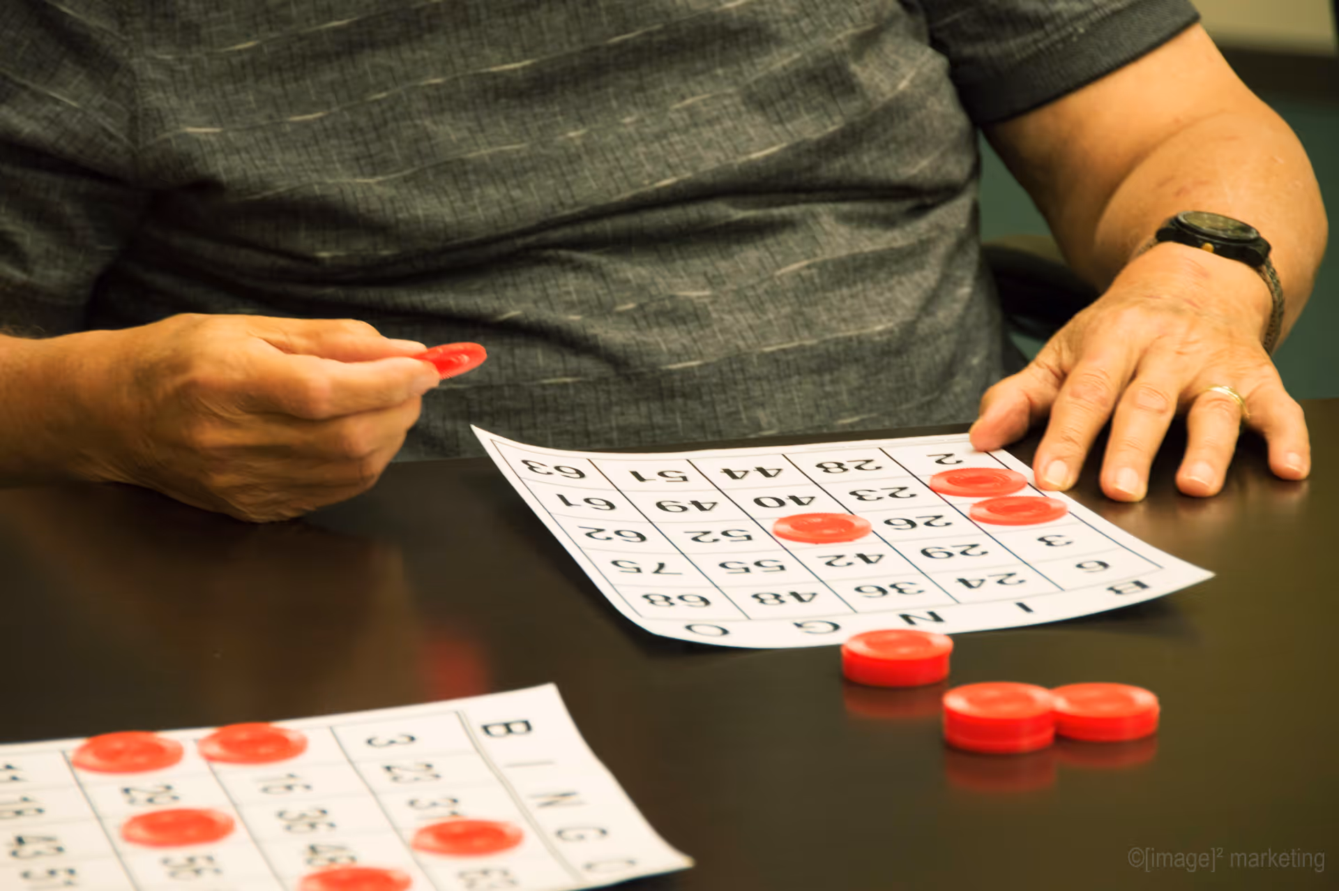 A person playing bingo, placing red markers on a bingo card on a table. Another bingo card with red markers is also visible on the table.
