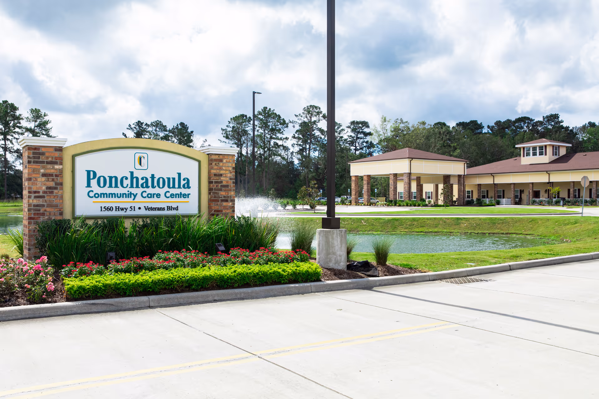 Exterior view of Ponchatoula Community Care Center showing the facility's sign with address 1560 Hwy 51 Veterans Blvd, a landscaped area with flowers and shrubs, a pond with a water fountain, and the building with a covered entrance under a partly cloudy sky.