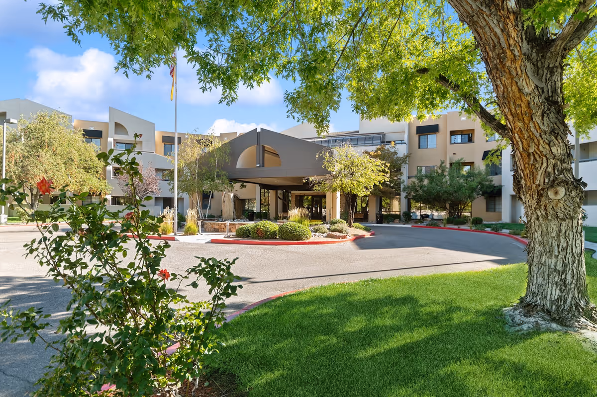 Exterior view of The Montebello on Academy senior living facility showing the main entrance with a covered drop-off area, surrounded by trees, bushes, and a well-maintained lawn under a clear blue sky.