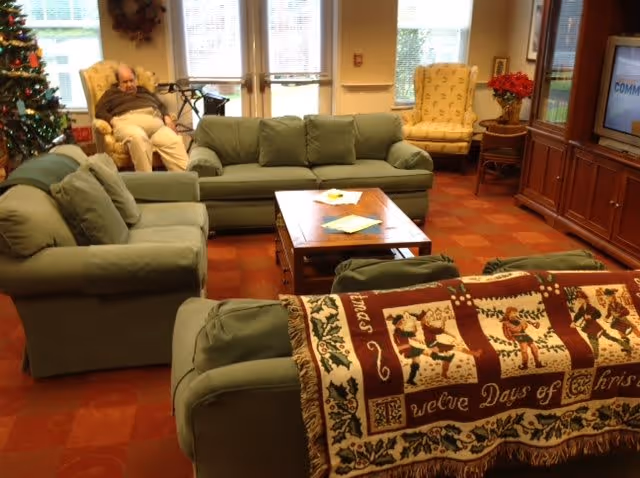 A cozy living room area in a senior living facility with green upholstered sofas and armchairs arranged around a wooden coffee table. A festive holiday blanket with Christmas-themed designs is draped over one sofa. In the background, a man is seated in a beige armchair near a decorated Christmas tree and a walker. The room has large windows with blinds, a wooden entertainment center with a TV, and warm lighting.
