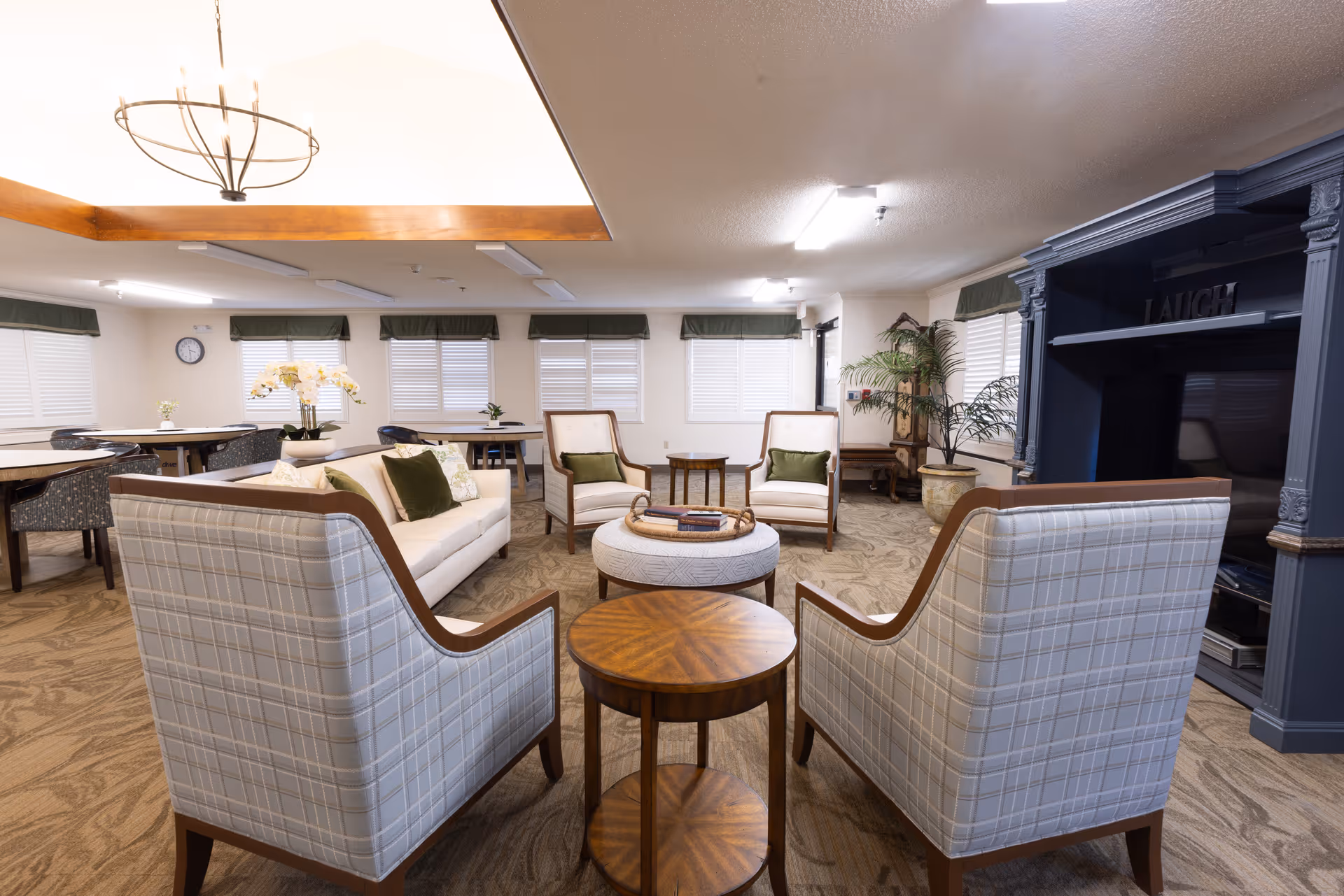 A spacious and well-lit common area with comfortable seating including two plaid armchairs, a white sofa with green cushions, and two wooden chairs with green cushions. There are round wooden tables and a large blue entertainment center with the word 'LAUGH' on the top shelf. The room has multiple windows with white blinds and green valances, beige carpet, and a chandelier hanging from the ceiling.
