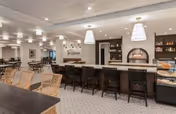 A spacious and well-lit dining area featuring a long bar with black bar stools, a wood-fired oven built into the wall, and multiple tables and chairs arranged for seating. The room has a neutral color palette with modern light fixtures hanging from the ceiling and decorative shelving on the walls.