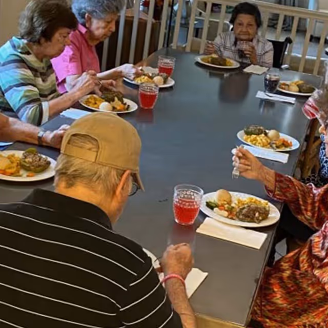 A group of elderly people sitting around a large rectangular table eating a meal together. Each person has a plate of food and a glass of red drink. The setting appears to be a communal dining area.