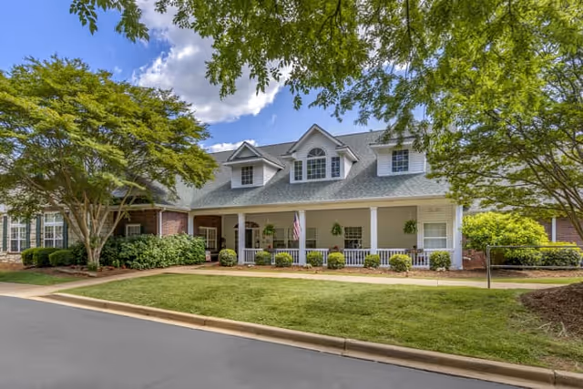 Front exterior of an assisted living building with a covered porch, dormer windows, columns, an American flag, and landscaped lawn framed by trees.