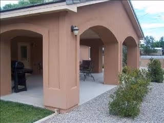 Covered outdoor patio area with arched openings, a barbecue grill, and outdoor seating. The patio is adjacent to a building with a tan stucco exterior and surrounded by gravel and some greenery.