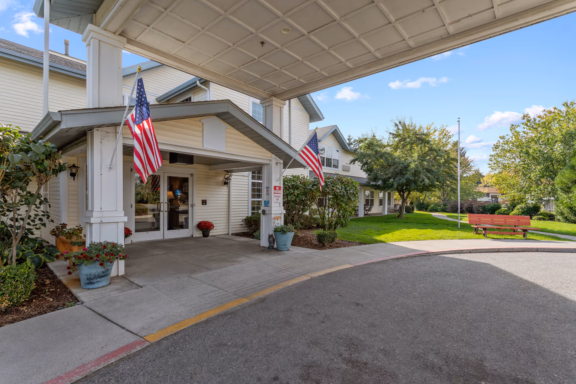 Covered entrance of a senior living building with American flags, potted plants, and a bench on a landscaped lawn.