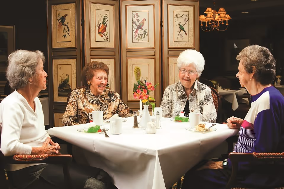 Four elderly women sitting around a dining table in a warmly lit room, enjoying conversation and dessert. The table is covered with a white tablecloth and has cups, plates with cake, and a small vase with flowers. Behind them is a decorative screen with bird illustrations.