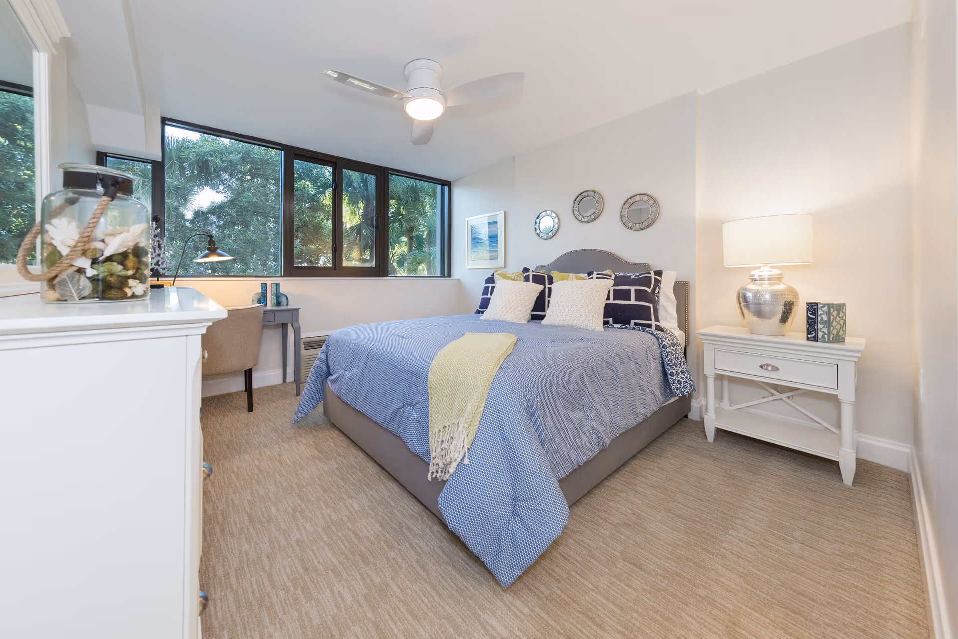A bright and cozy bedroom with a large bed covered in blue bedding and multiple pillows. There is a white nightstand with a silver lamp and a small decorative box on the right side of the bed. On the left side, there is a white dresser with a glass jar containing decorative items. A small desk with a chair and a lamp is positioned under a set of large windows showing green trees outside. The walls are light-colored, and the floor is carpeted.
