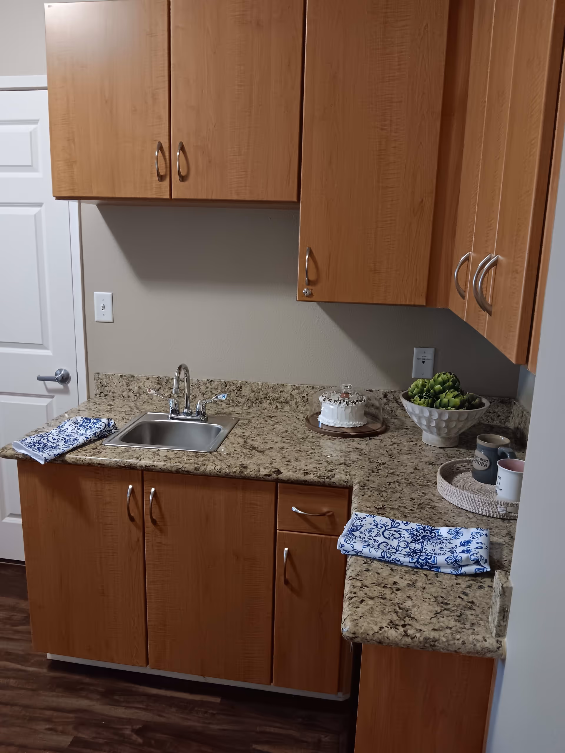 A small kitchen area with wooden cabinets and a granite countertop. There is a stainless steel sink with a faucet, two blue and white patterned cloths on the counter, a small white cake under a glass dome, a bowl of artichokes, and a tray with two mugs. The floor is dark wood, and a white door is visible in the background.