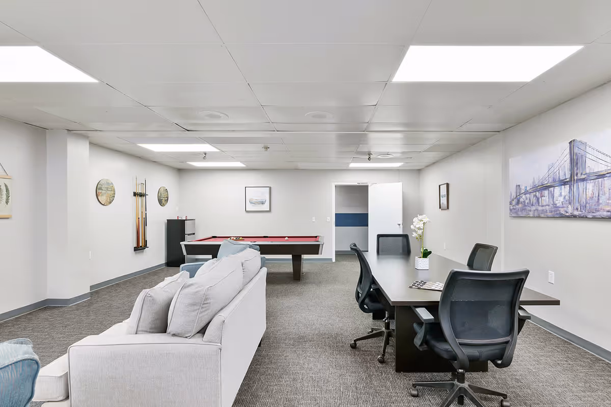 Open communal recreation room with sofas, a pool table, and a long table with office chairs under fluorescent ceiling lights.