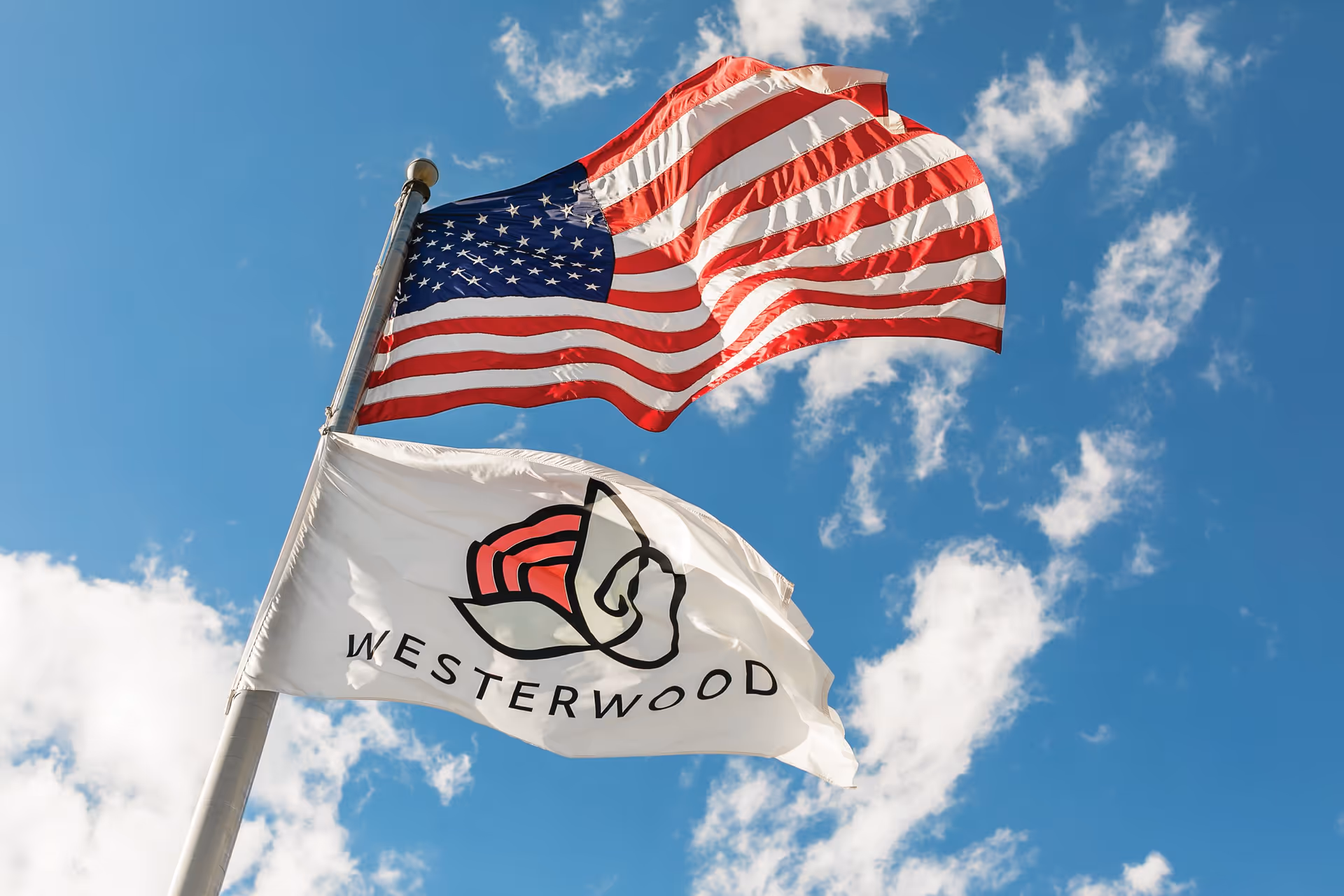 Two flags flying on a flagpole against a blue sky with scattered clouds. The top flag is the United States flag, and the bottom flag is white with a stylized red and black flower logo and the word 'WESTERWOOD' written below it.