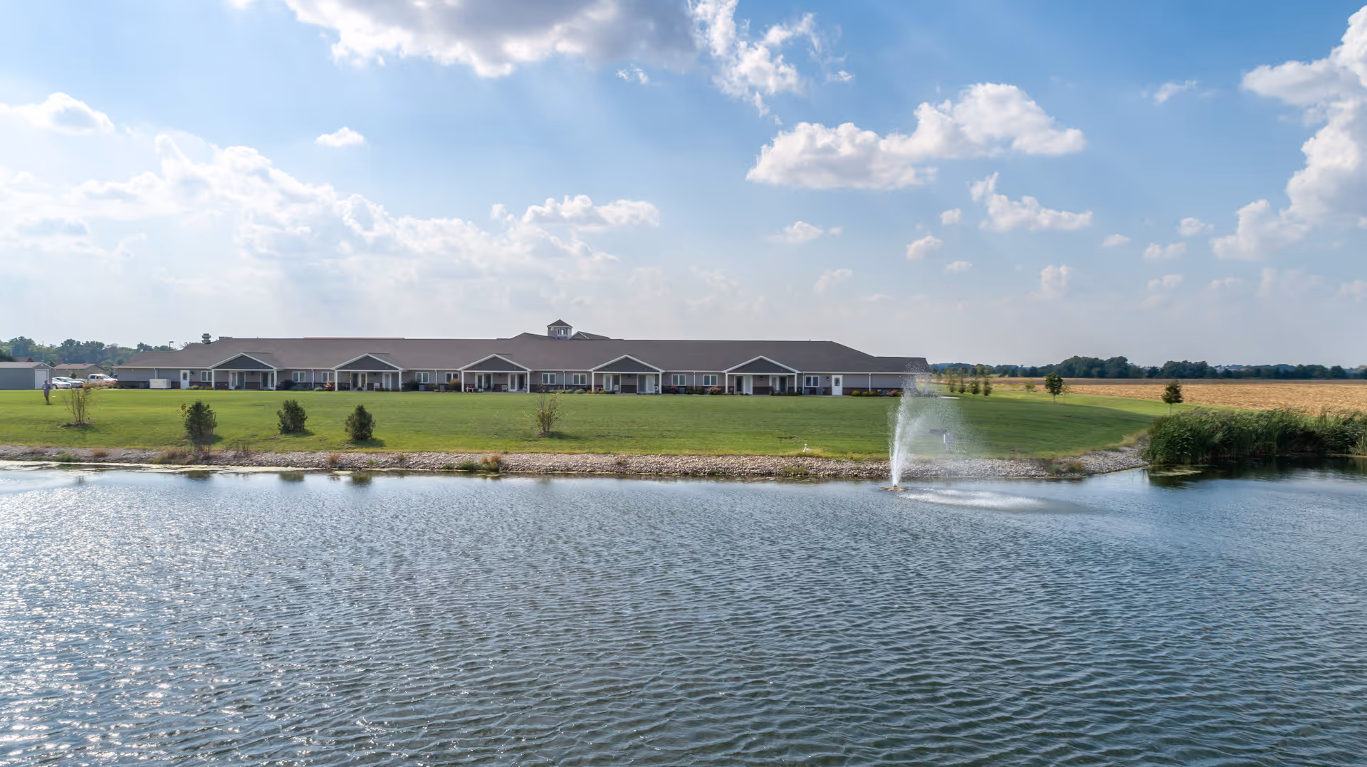 Single-story memory care building with covered porches facing a pond and a fountain under a partly cloudy sky.