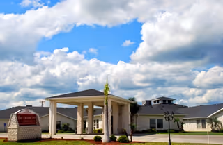 Front entrance of Rio Grande City Nursing and Rehabilitation Center with a covered portico, landscaped grounds, and a sign beneath a partly cloudy sky.