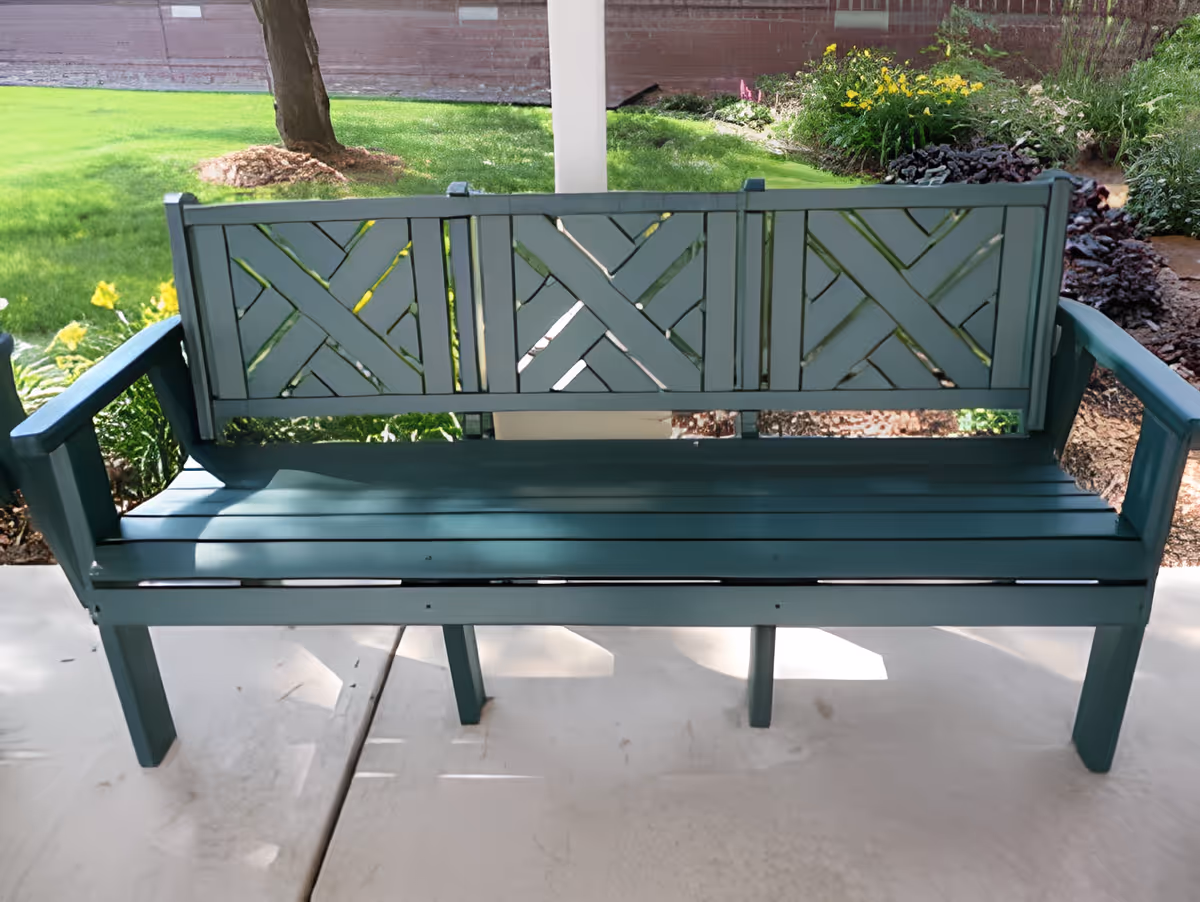 A teal wooden bench on a covered porch with lawn and flower beds visible behind it.