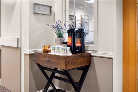 A small wooden table with a dark finish and crossed legs holds two black coffee dispensers, a tray with cups, and a vase with purple flowers. The table is placed against a beige and white wall with a window and a framed sign that reads 'blessed'.