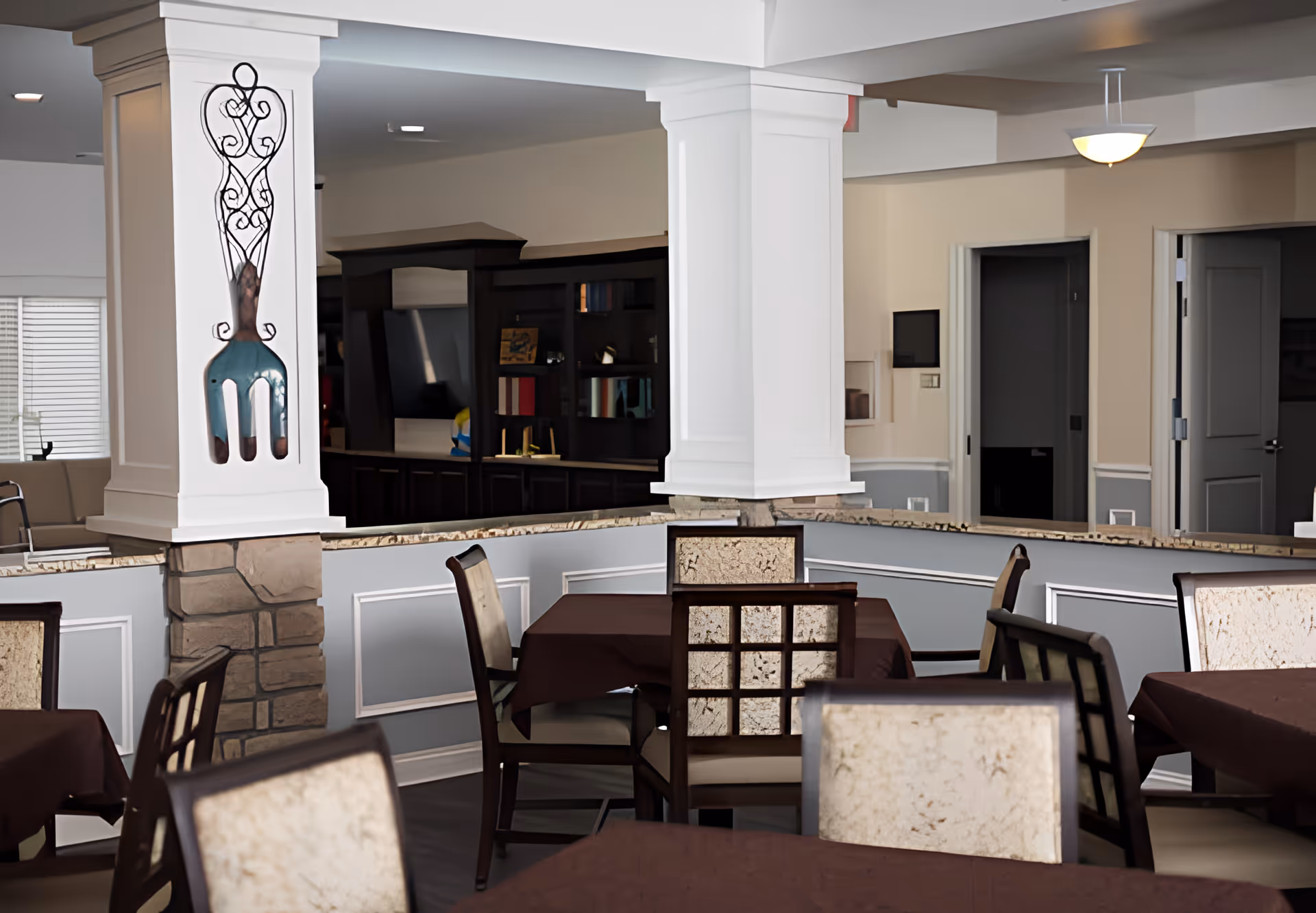 Interior view of a dining area with several tables covered in dark brown tablecloths and chairs with light-colored cushioned backs and seats. The room features white columns with decorative stone bases and a large decorative fork wall art on one column. In the background, there is a dark wooden cabinet with shelves and a TV, and several doorways leading to other rooms. The lighting is soft with ceiling lights and a hanging lamp.